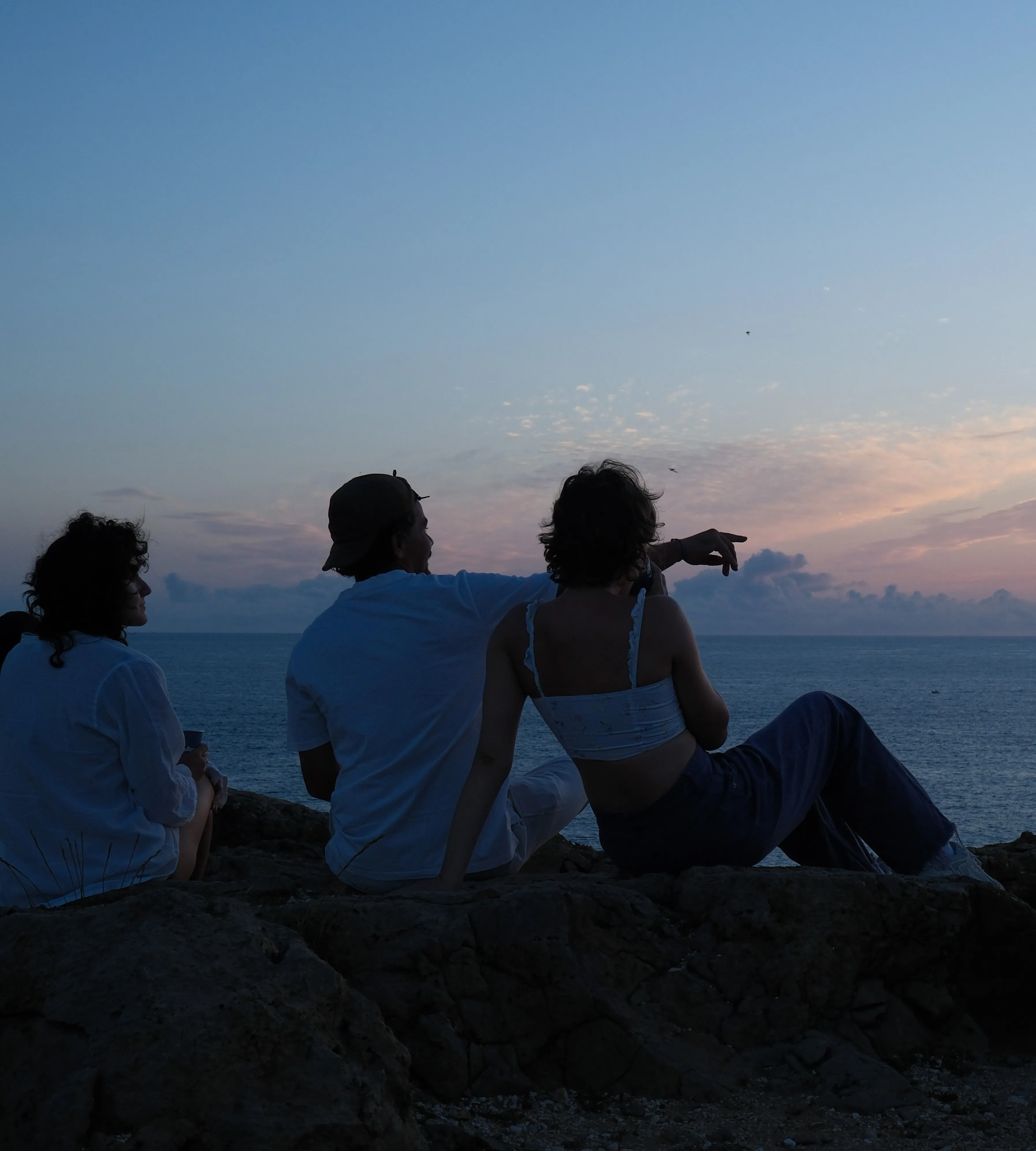 Three people sitting on the rocks at dusk, looking out to sea